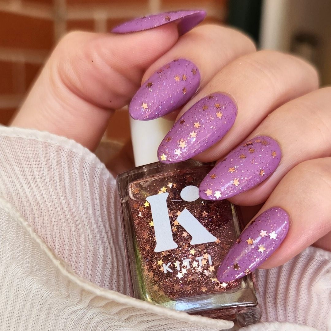 Hand with purple glittery nails holding a bottle of nail polish with a visible brand logo.