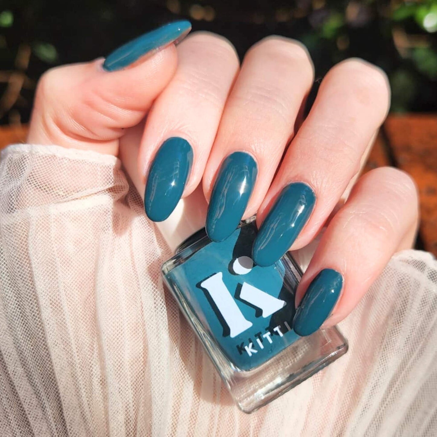Hand with long almond nails painted deep teal, holding Kitti Nails In Too Deep bottle