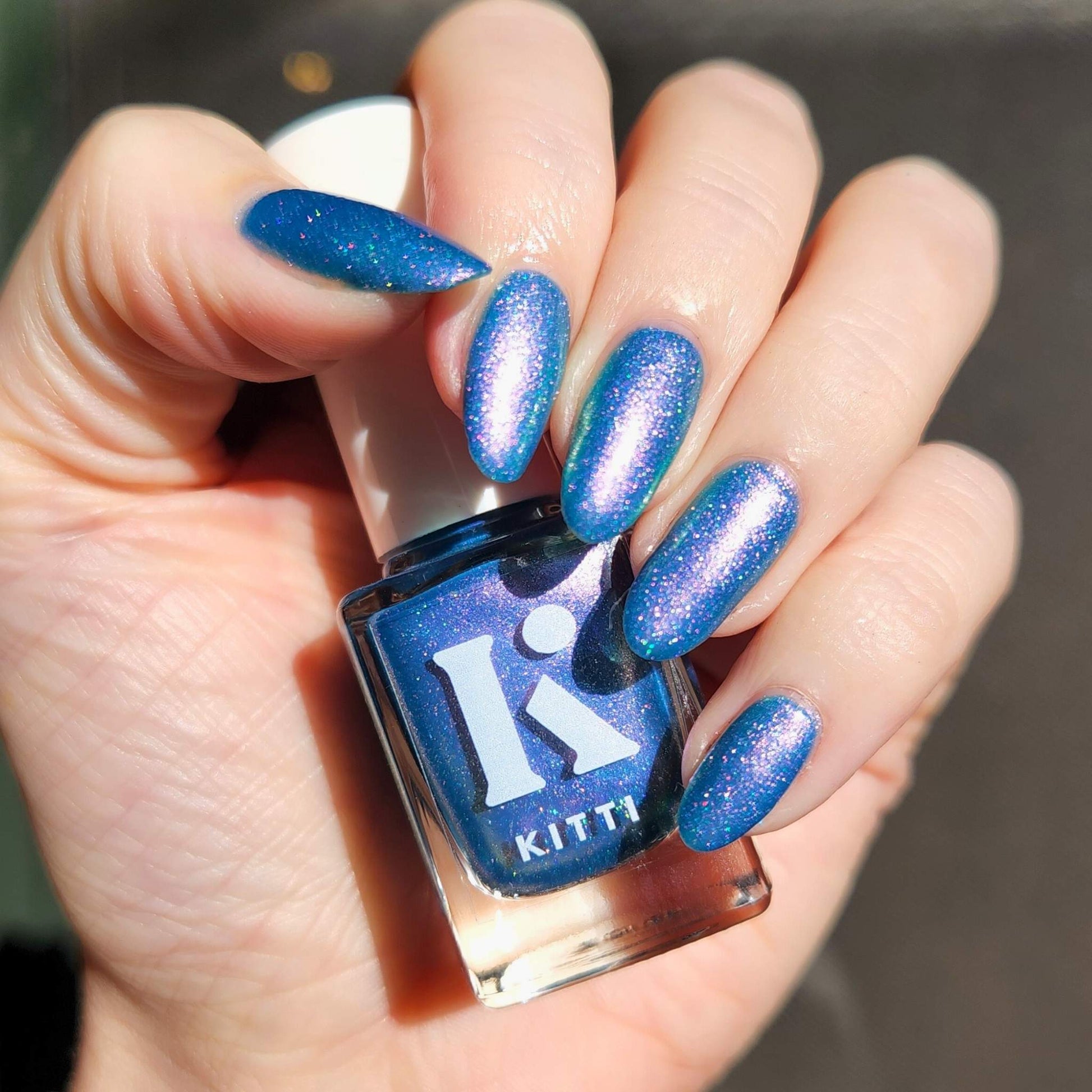 Hand with blue glittery nails holding a bottle of Kitti nail polish.