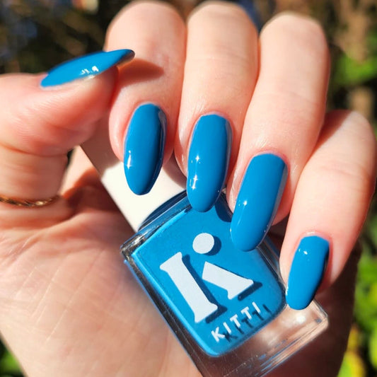 Hand with long almond nails painted bright blue, holding Kitti Nails Ocean Drive bottle